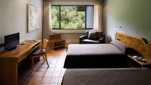 A hotel room with two beds, a desk with a TV, a chair, and a window with a view of trees. - As Hoteles Altube (Zuia)