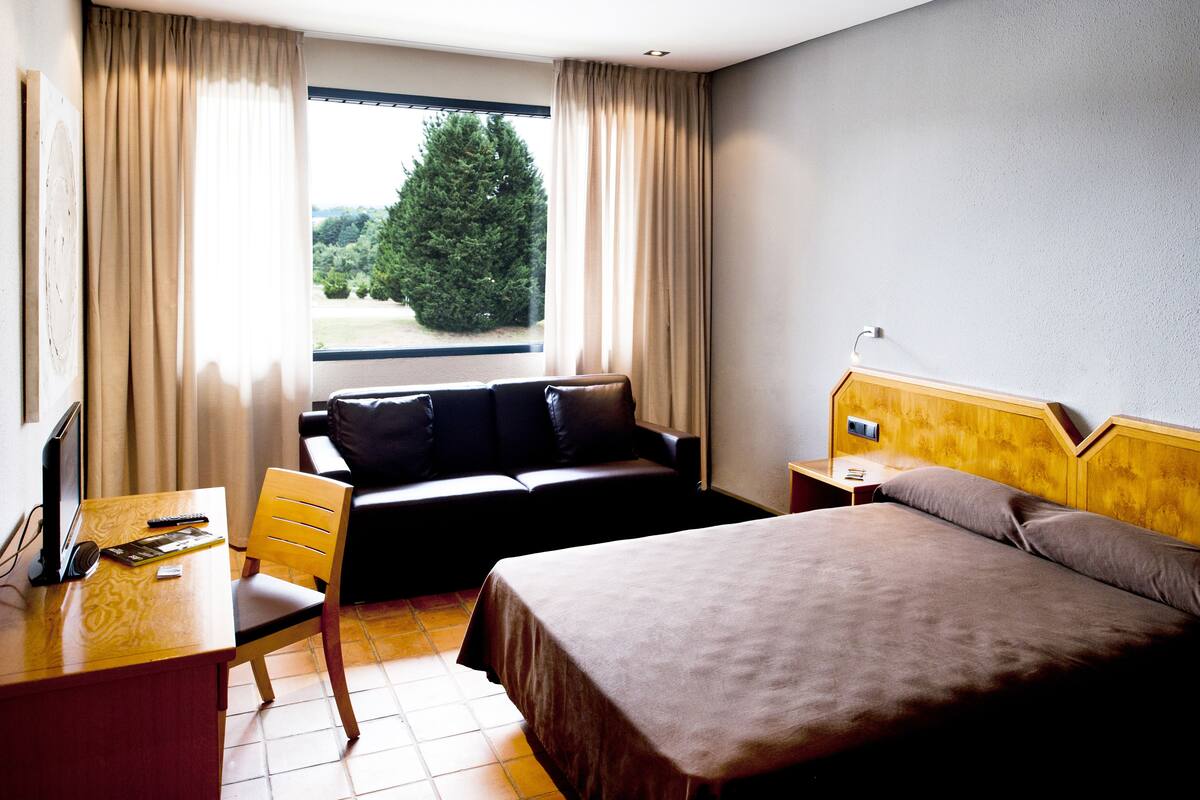 A hotel room with a bed, a desk with a computer, a chair, and a window with curtains.