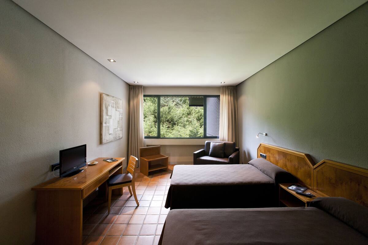 A hotel room with two beds, a desk with a computer, a chair, and a window with a view of trees.