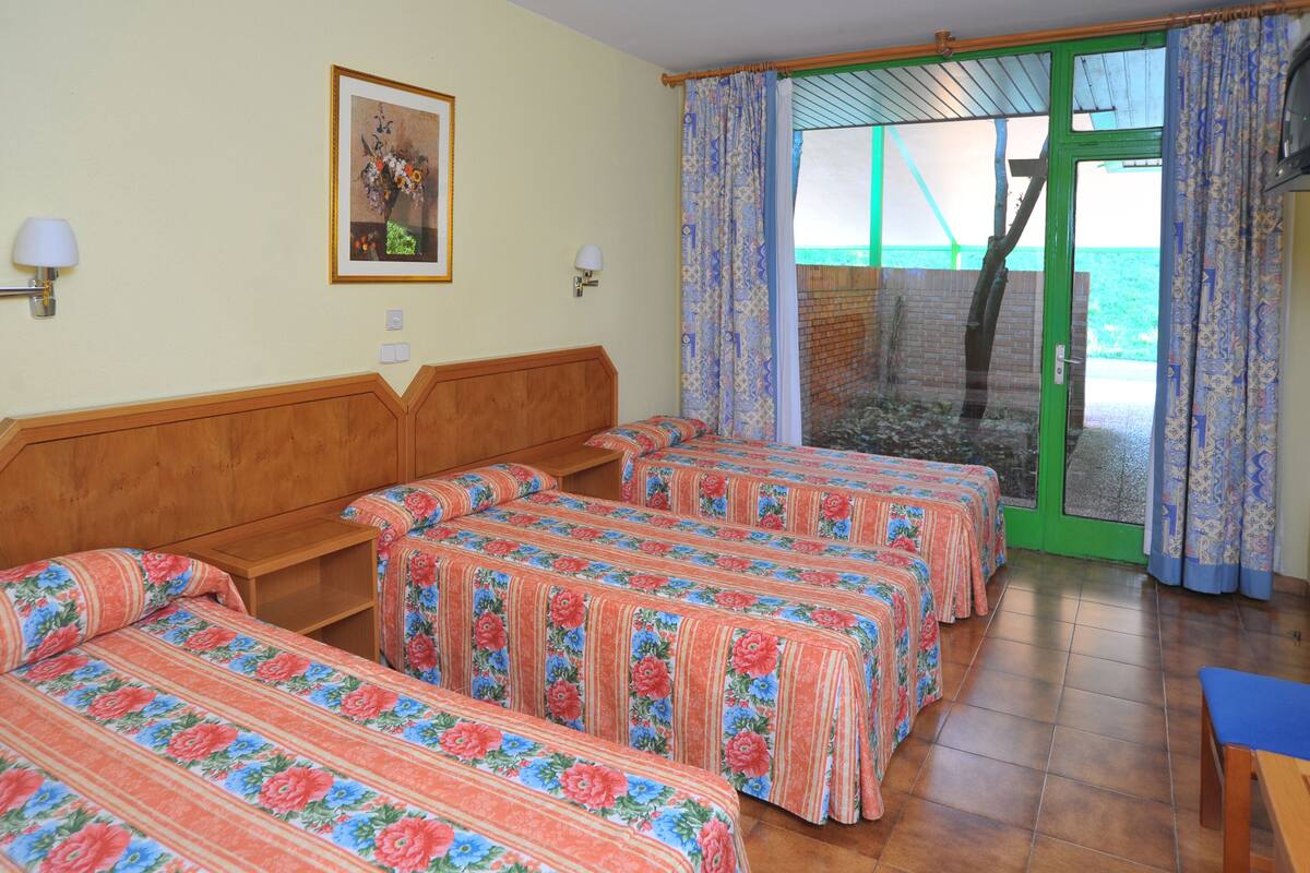 A hotel room with four single beds, a wooden headboard, a small table, and a view of the sea through a glass door.