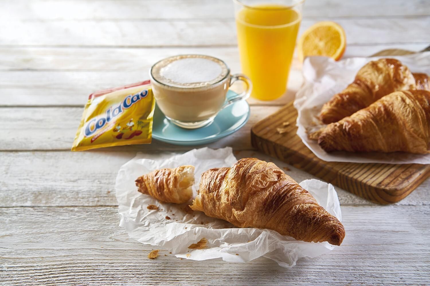 Croissants, a cup of coffee, a glass of orange juice, and a packet of sugar on a wooden table.