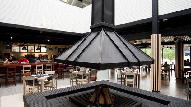 A modern indoor space with a central black grill area, surrounded by tables and chairs, under a large glass ceiling. - As Hoteles Altube (Zuia)