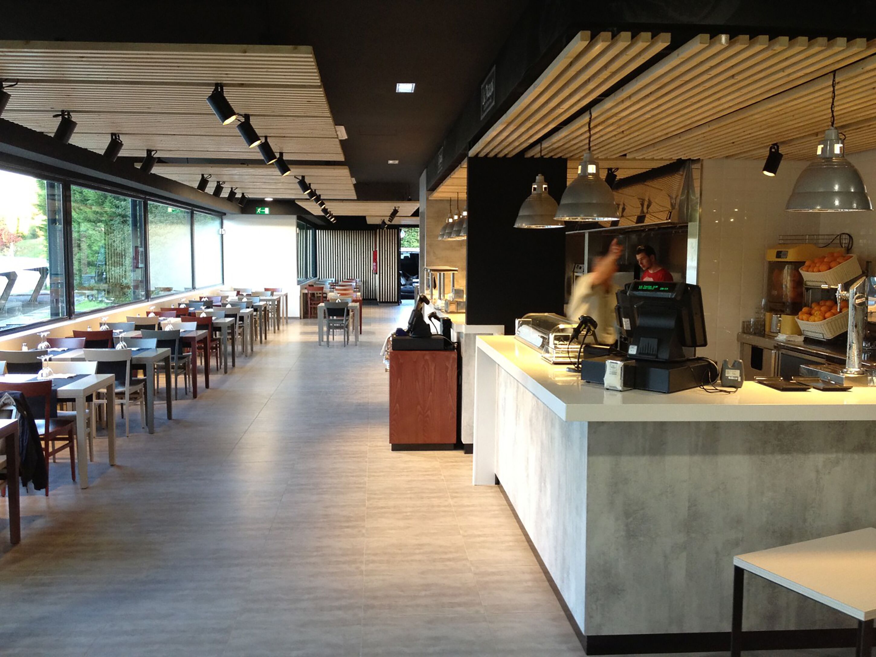 A spacious cafe with long tables and chairs, a counter with a coffee machine, and large windows providing natural light.