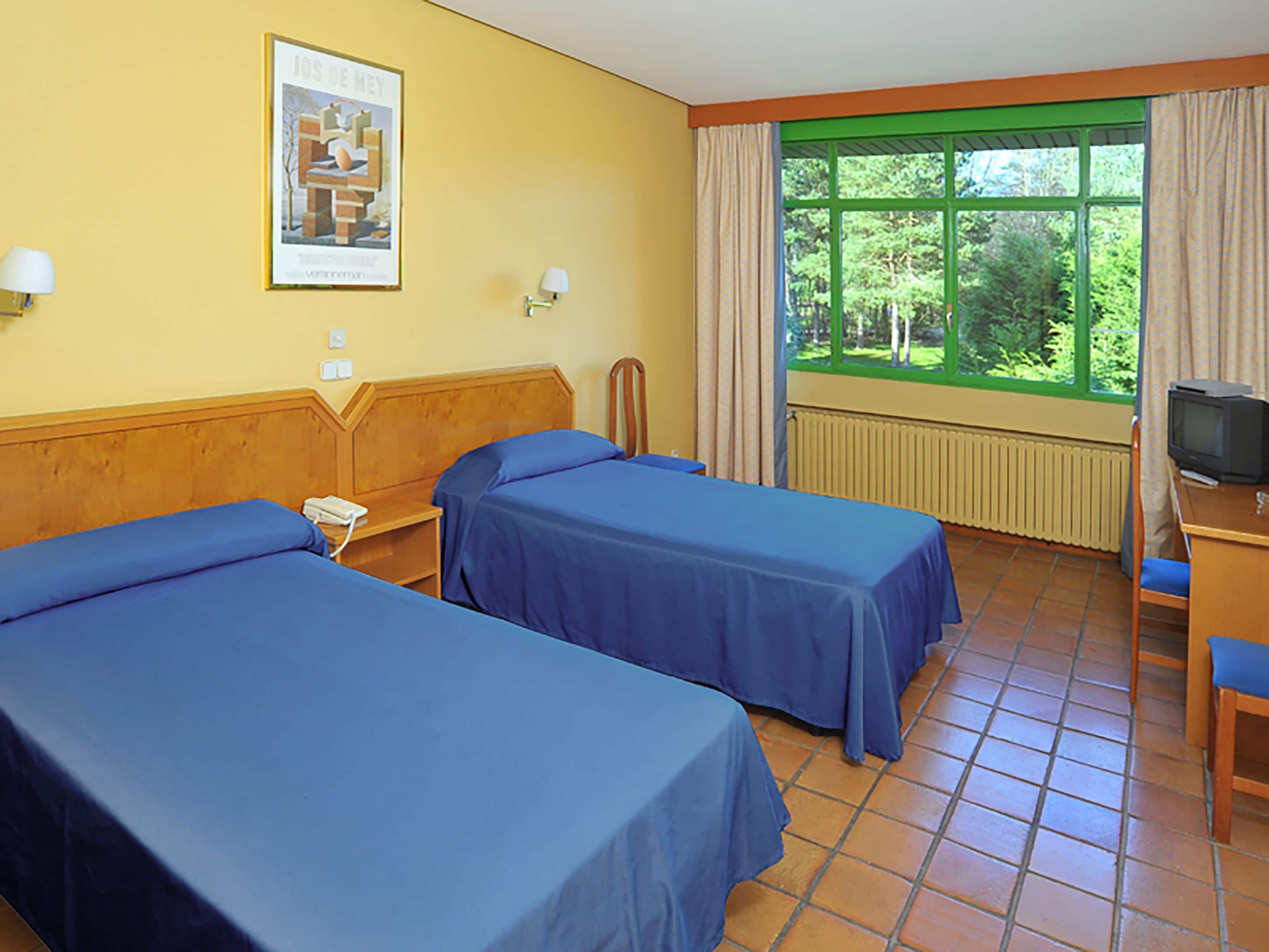 A hotel room with two single beds, a small desk with a television, a window with curtains, and a poster on the wall.