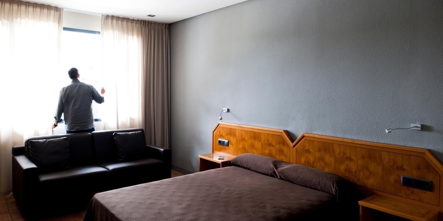 Une chambre d’hôtel avec un lit, une tête de lit en bois, une table de chevet, une lampe, un canapé en cuir noir et une fenêtre avec des rideaux.