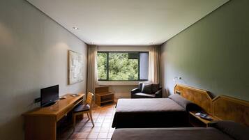 A hotel room with two beds, a desk with a computer, a chair, a window with a view of greenery, and a wall-mounted TV.