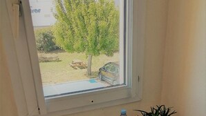 Une petite table sur laquelle se trouve un plateau avec un croissant, une pomme, une tasse de café et une bouteille d’eau. Une plante en pot est posée sur la table, et une fenêtre donne sur un arbre et une voiture.
