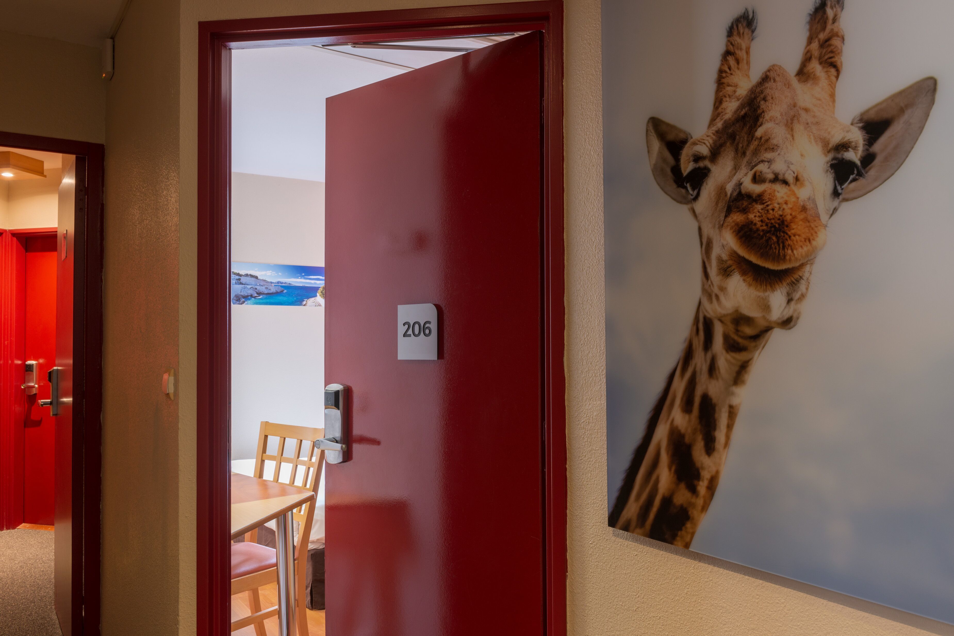 Una camera d'hotel con una porta rossa, il numero '206' sulla parete e un murale raffigurante una giraffa.