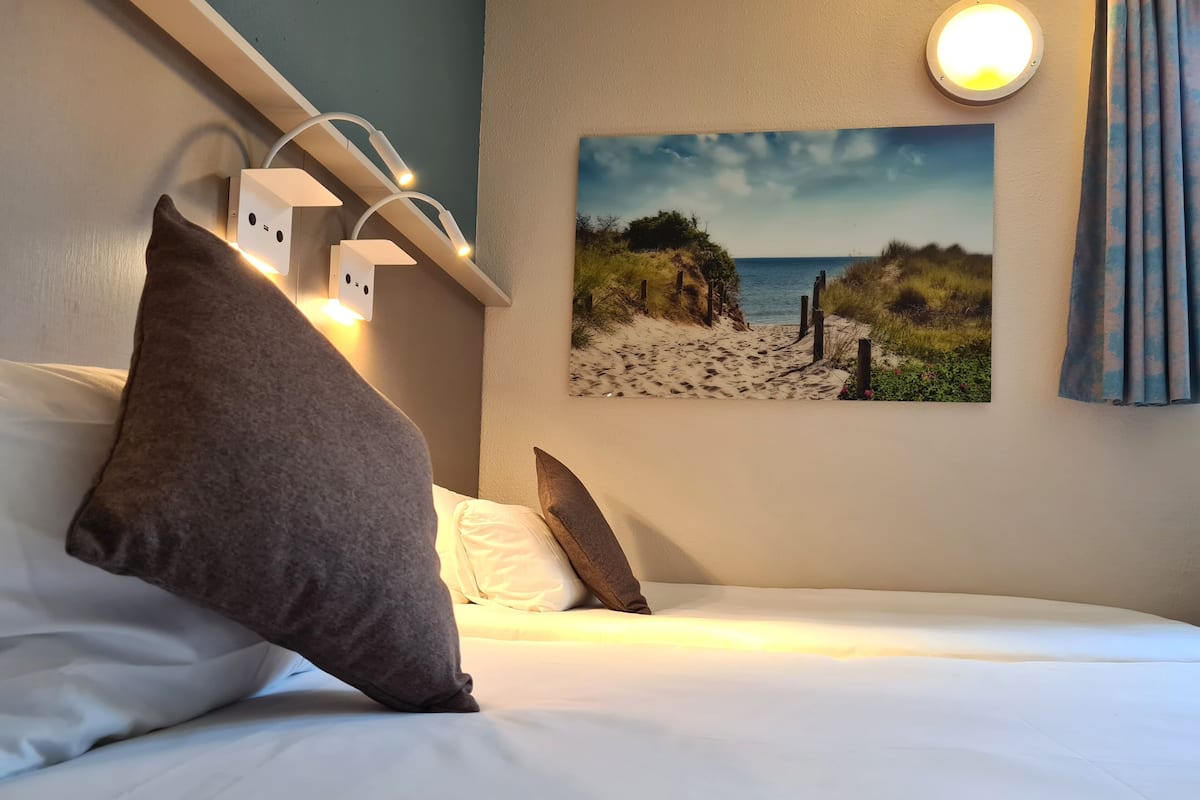 Habitació d'hotel amb llit, almoines, llum penjant a la paret, quadre d'una platja i finestra amb cortines.