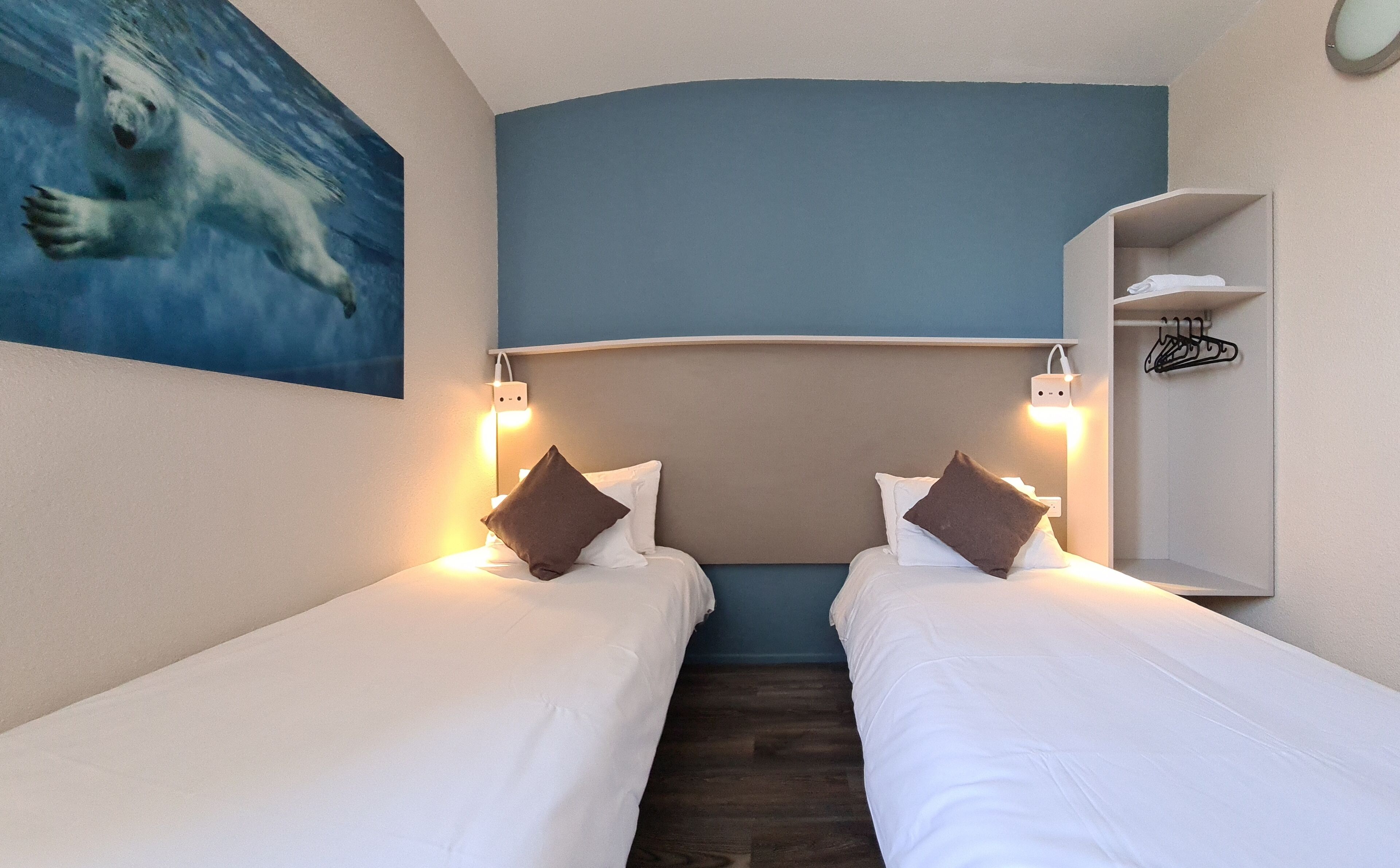 A hotel room with two single beds, a headboard, bedside lamps, a painting of a polar bear, and a small shelf with a hanger.