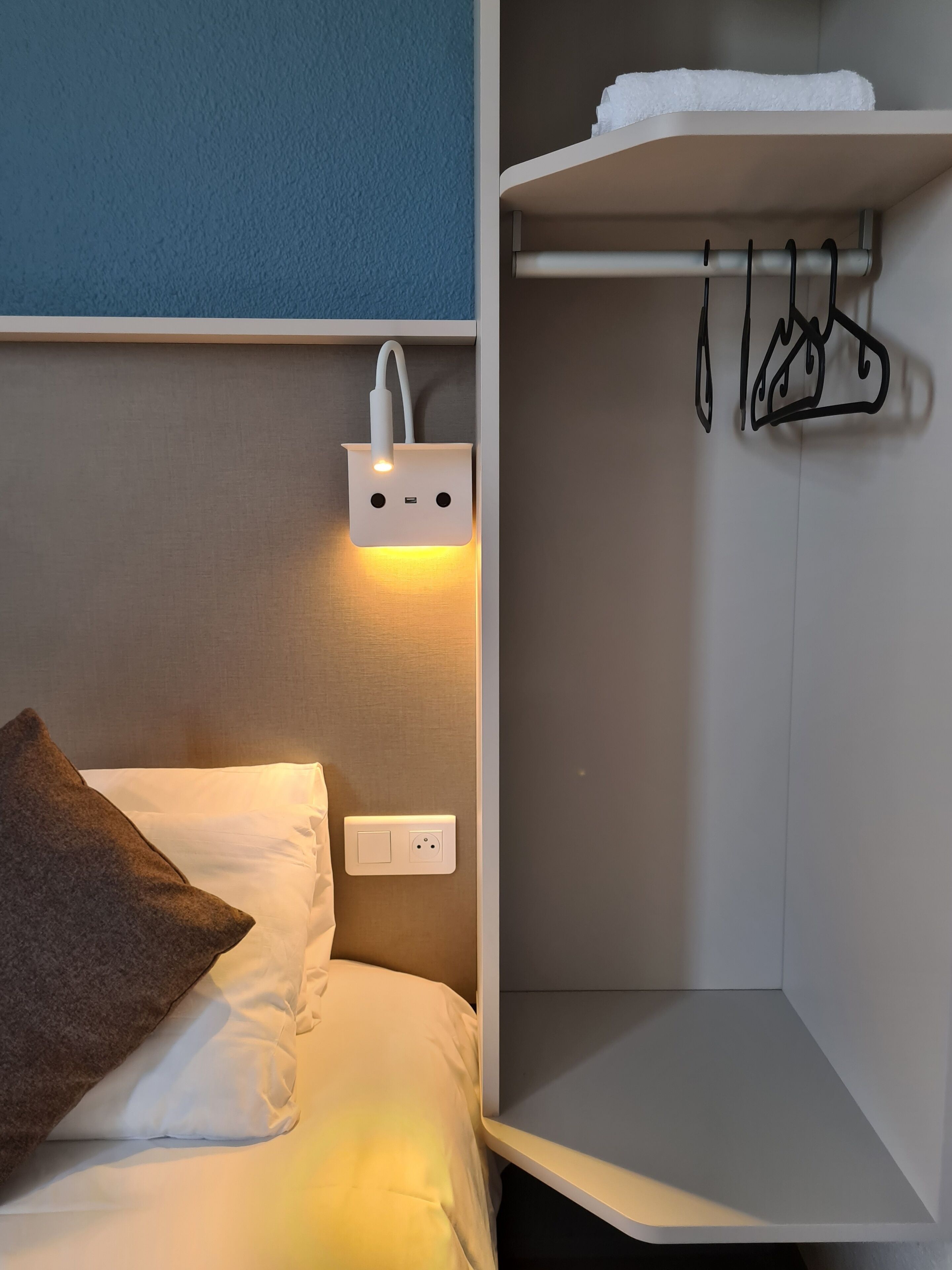 A hotel room with a bed, a wall-mounted light, a coat rack, and a shelf.
