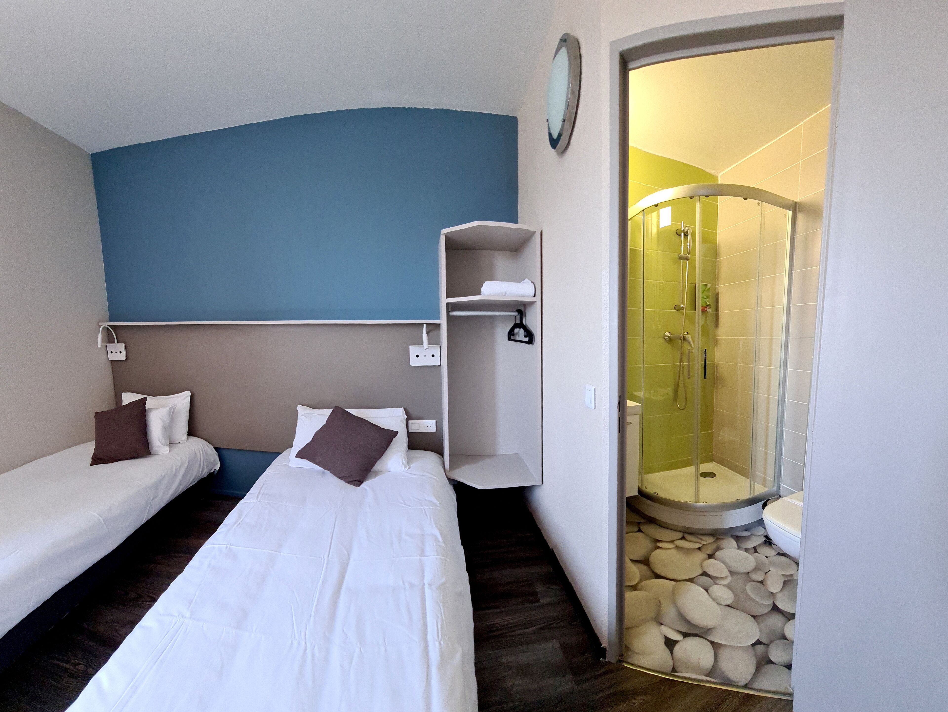 Habitación de hotel con dos camas individuales, baño con ducha de vidrio y una pared de acento azul.