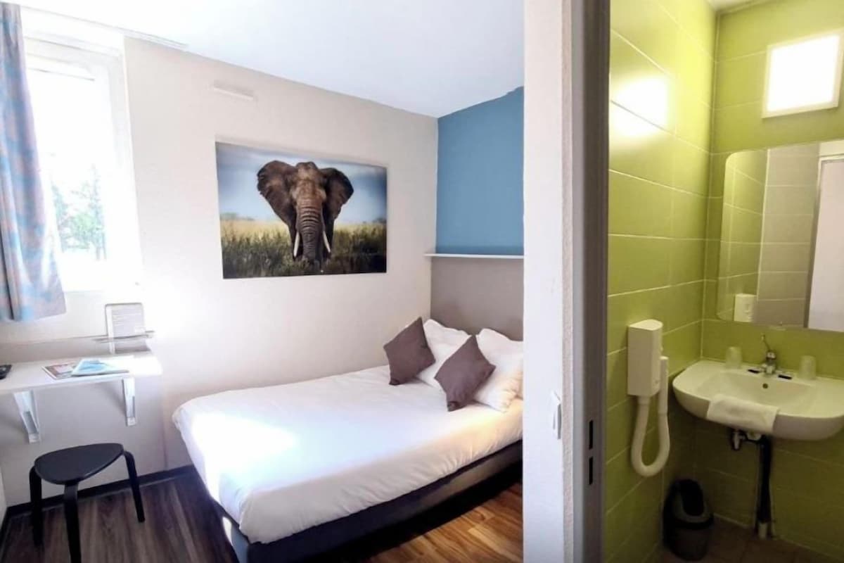 Ein Schlafzimmer mit Bett, Schreibtisch, Stuhl, einem Elefantenbild und einem Badezimmer mit Waschbecken und Spiegel.