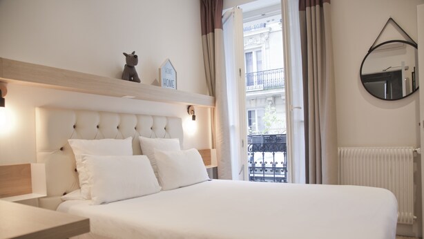 A neatly made bed with white bedding, a bedside table with a lamp, a small shelf with decorative items, and a view of a balcony with a railing. - Alexandrine Opera (Paris)