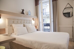 Ein ordentlich bezogenes Bett mit weißer Bettwäsche, ein Nachttisch mit Lampe, eine kleine Ablagefläche mit Dekoration und Blick auf einen Balkon mit Geländer.