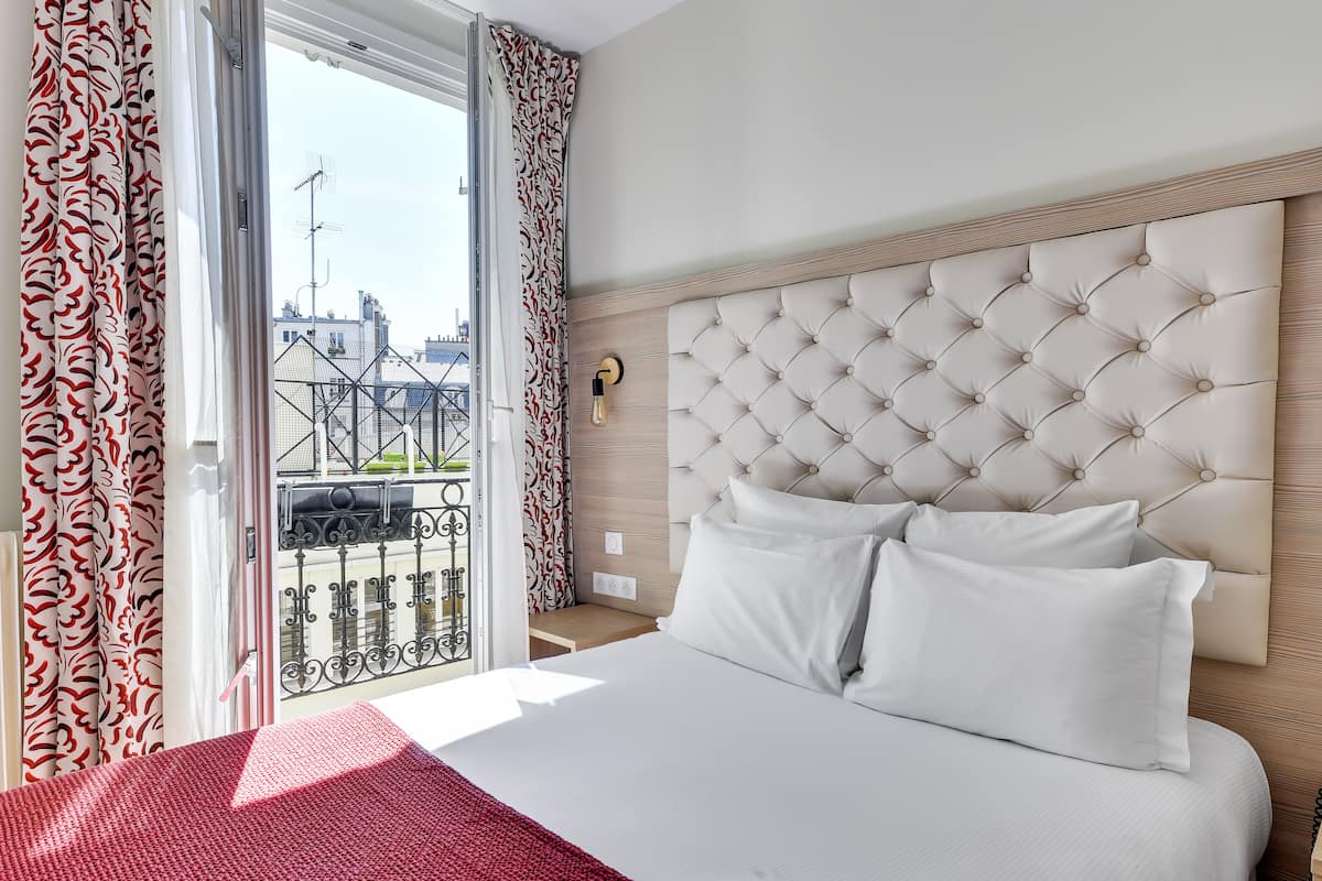 A hotel room with a large bed, a tufted headboard, a red throw blanket, and a view of the city through a balcony.