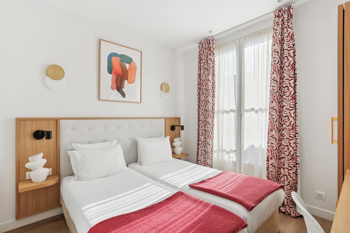 A hotel room with two beds, a large window with patterned curtains, a wall-mounted artwork, and a wooden nightstand with decorative items.