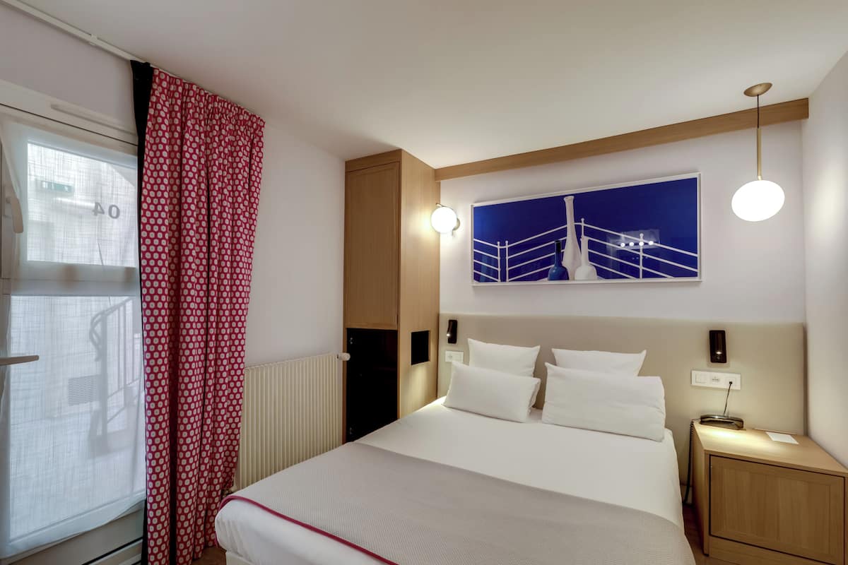 A hotel room with a bed, bedside table, wall-mounted light, and a painting above the bed.