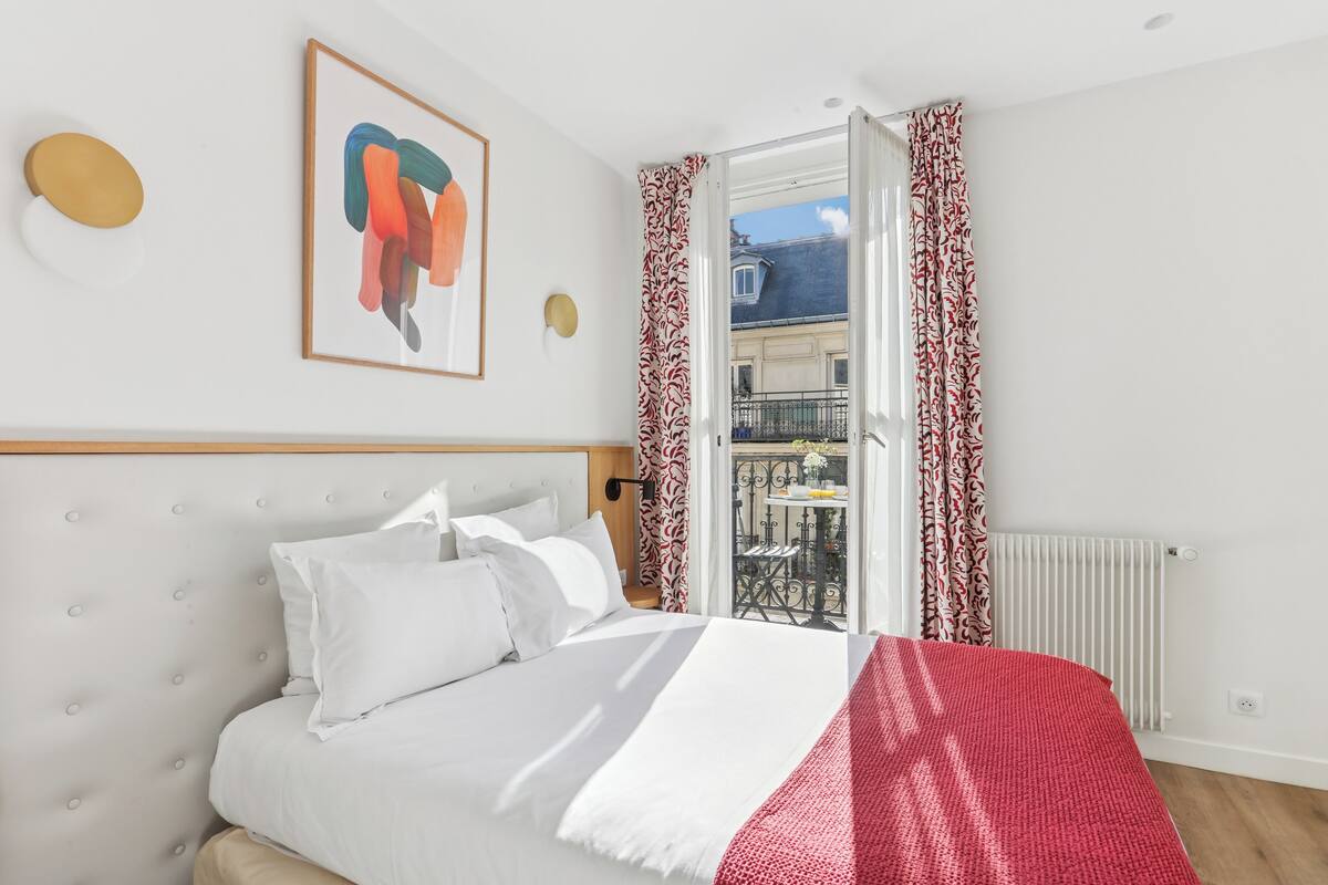 A neatly made bed with white bedding, a red blanket, and a headboard with a tufted design. There is a framed abstract painting on the wall, and a window with patterned curtains.