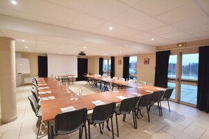 Een vergaderzaal met een lange tafel, stoelen, een projectiescherm en grote ramen met gordijnen.