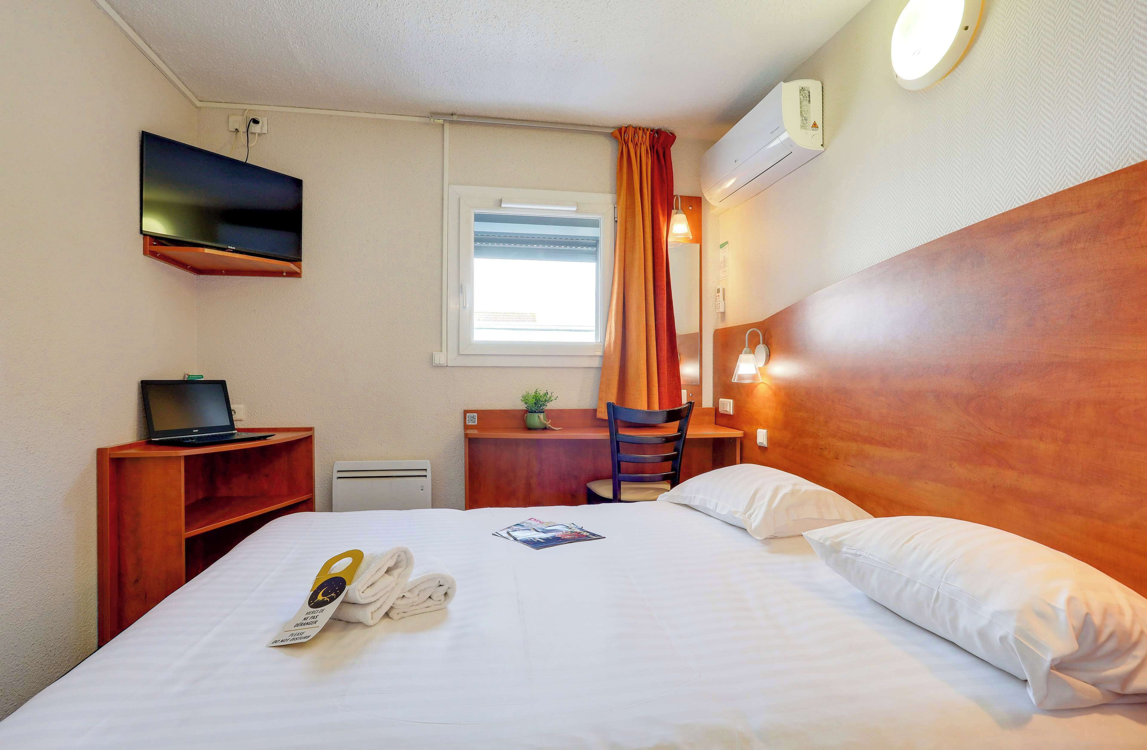 Una camera d'albergo con un letto, una scrivania con un portatile, una sedia, una TV a parete e una finestra con tende.