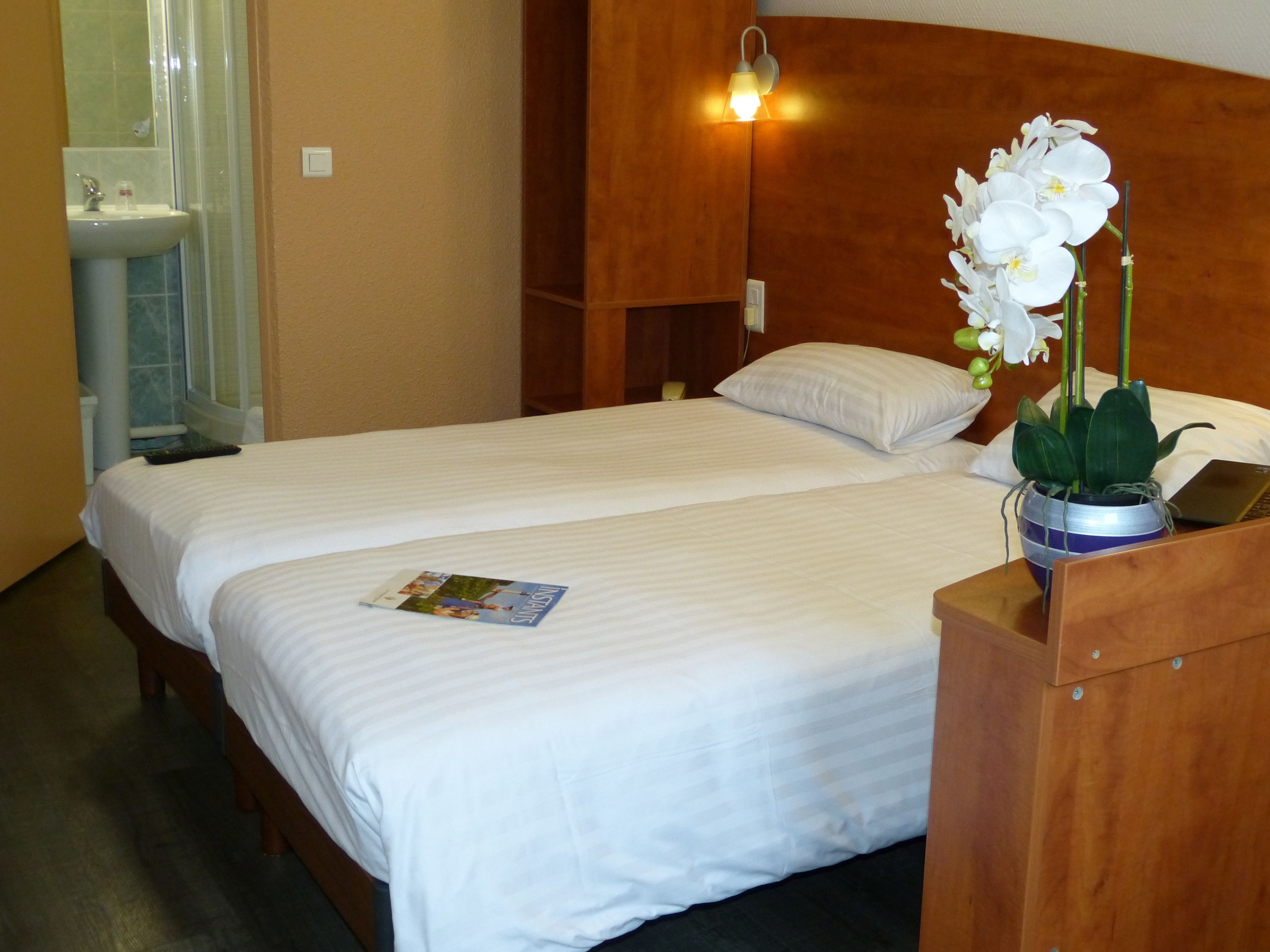 Habitación de hotel con dos camas, cabecero de madera, mesita de noche con un ordenador portátil, orquídea en maceta y una revista sobre una de las camas.