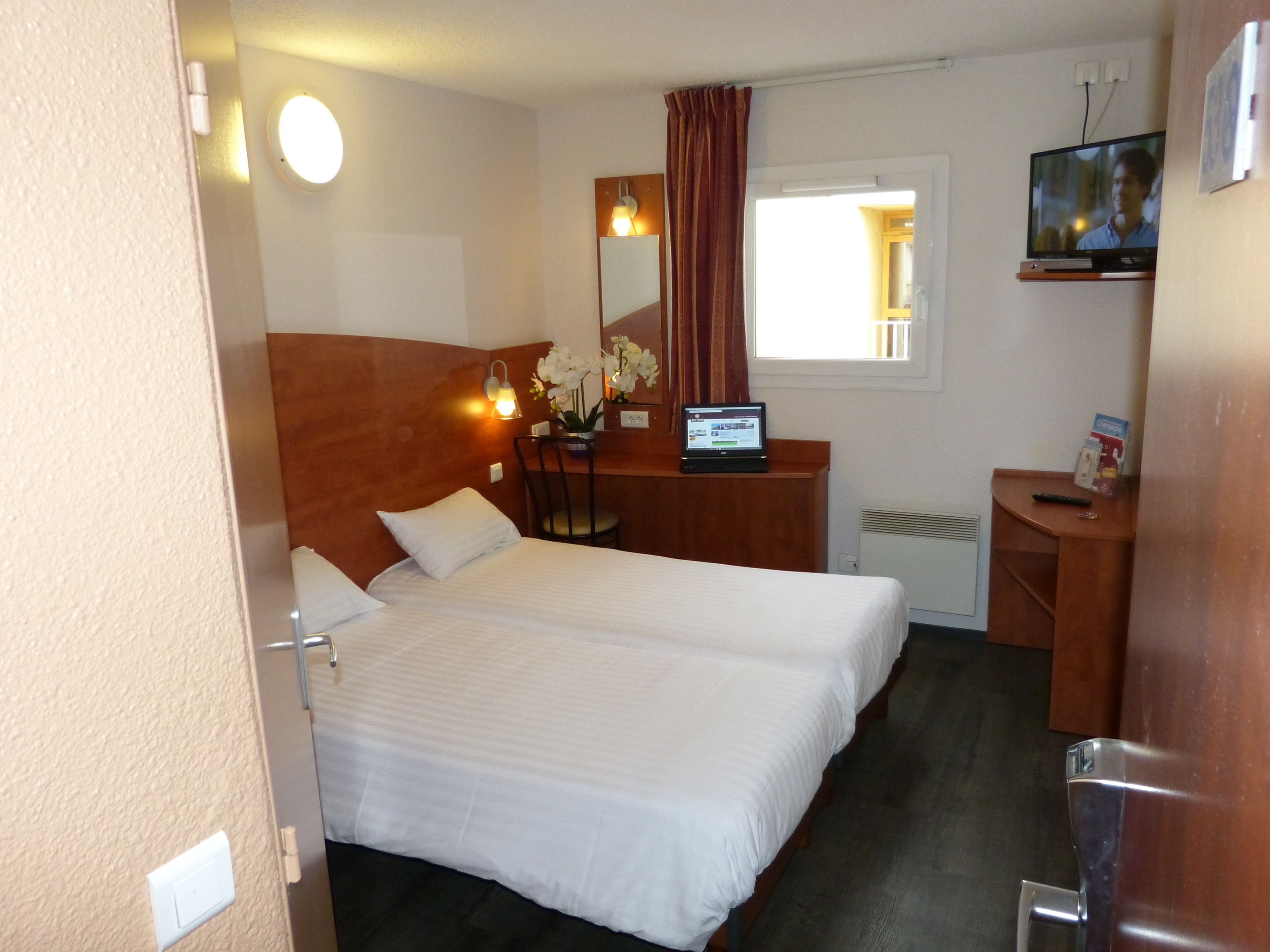 A hotel room with two beds, a desk with a laptop, a TV, and a window with curtains.