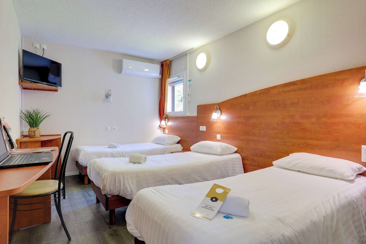 Habitación de hotel con dos camas individuales, un escritorio con una computadora portátil, una silla, un televisor montado en la pared y una ventana con cortinas.