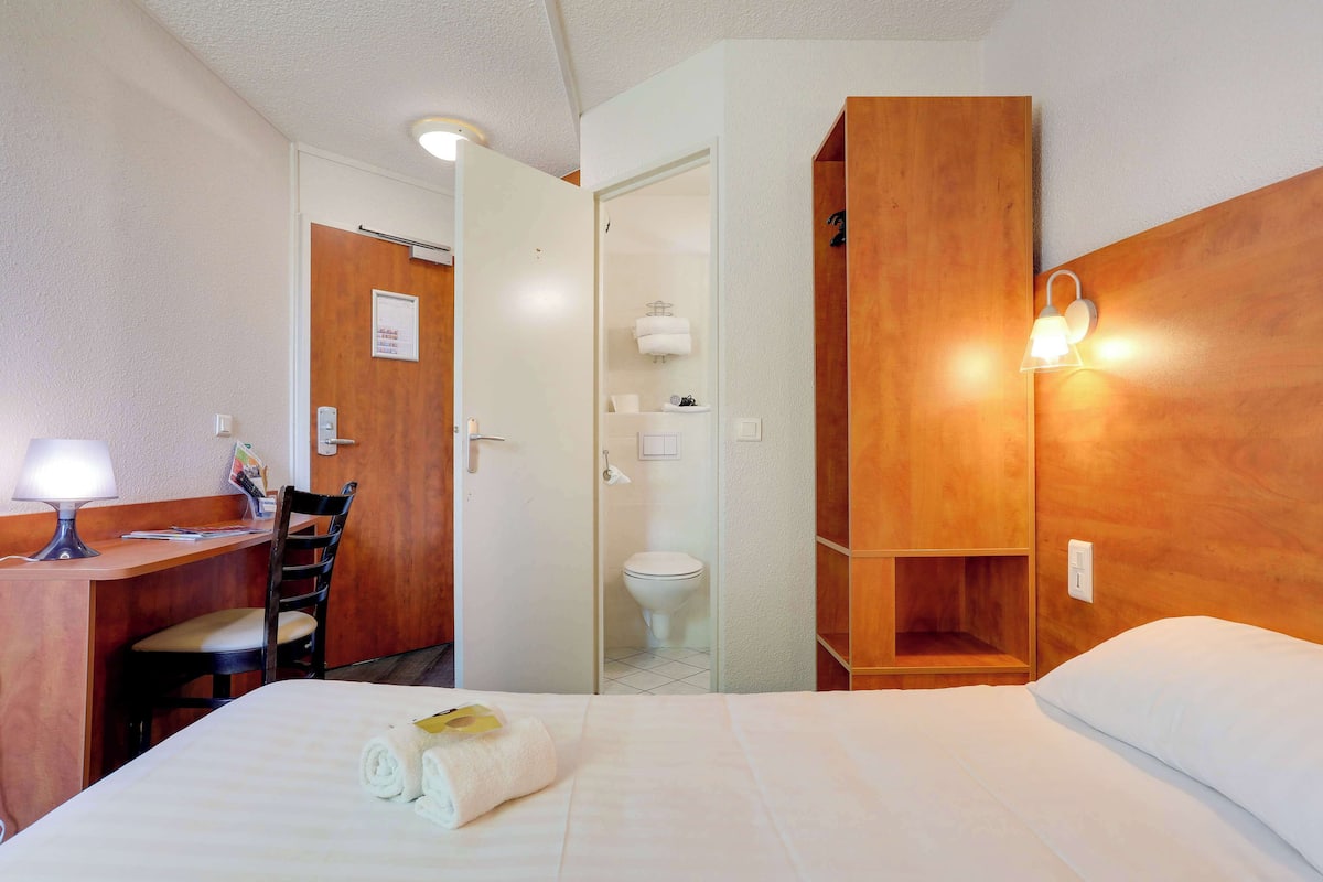 Habitación de hotel con cama, un escritorio y silla, baño con inodoro y lavamanos, y estante de madera.