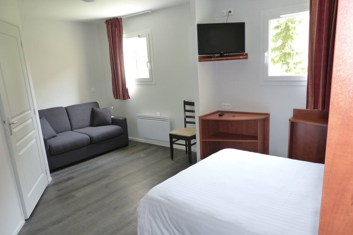 Un dormitorio con cama, sofá gris, escritorio de madera, televisor montado en la pared y ventana con cortinas.