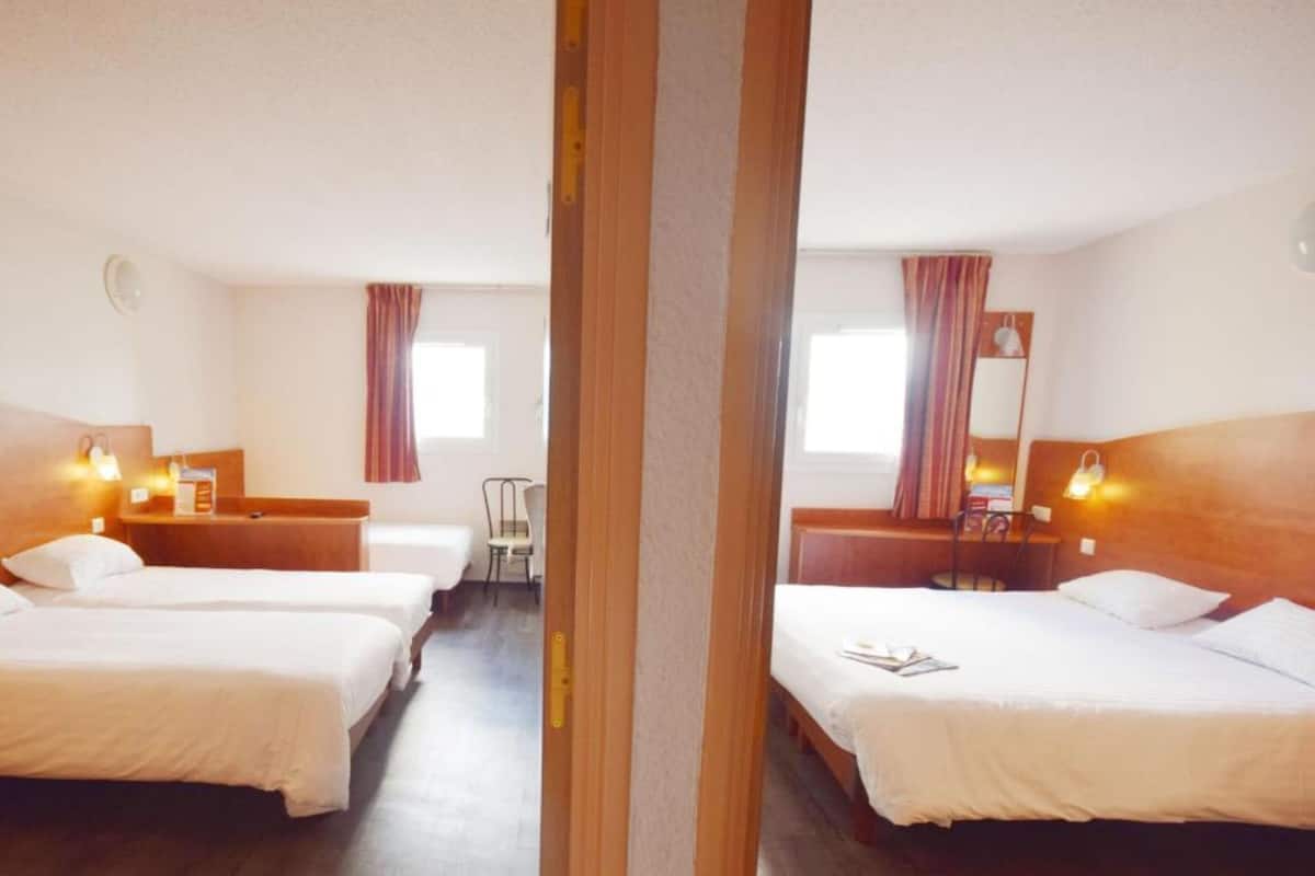 Habitación de hotel con dos camas individuales, cabecera de madera, escritorio pequeño con silla, ventana con cortinas y un espejo en la pared.