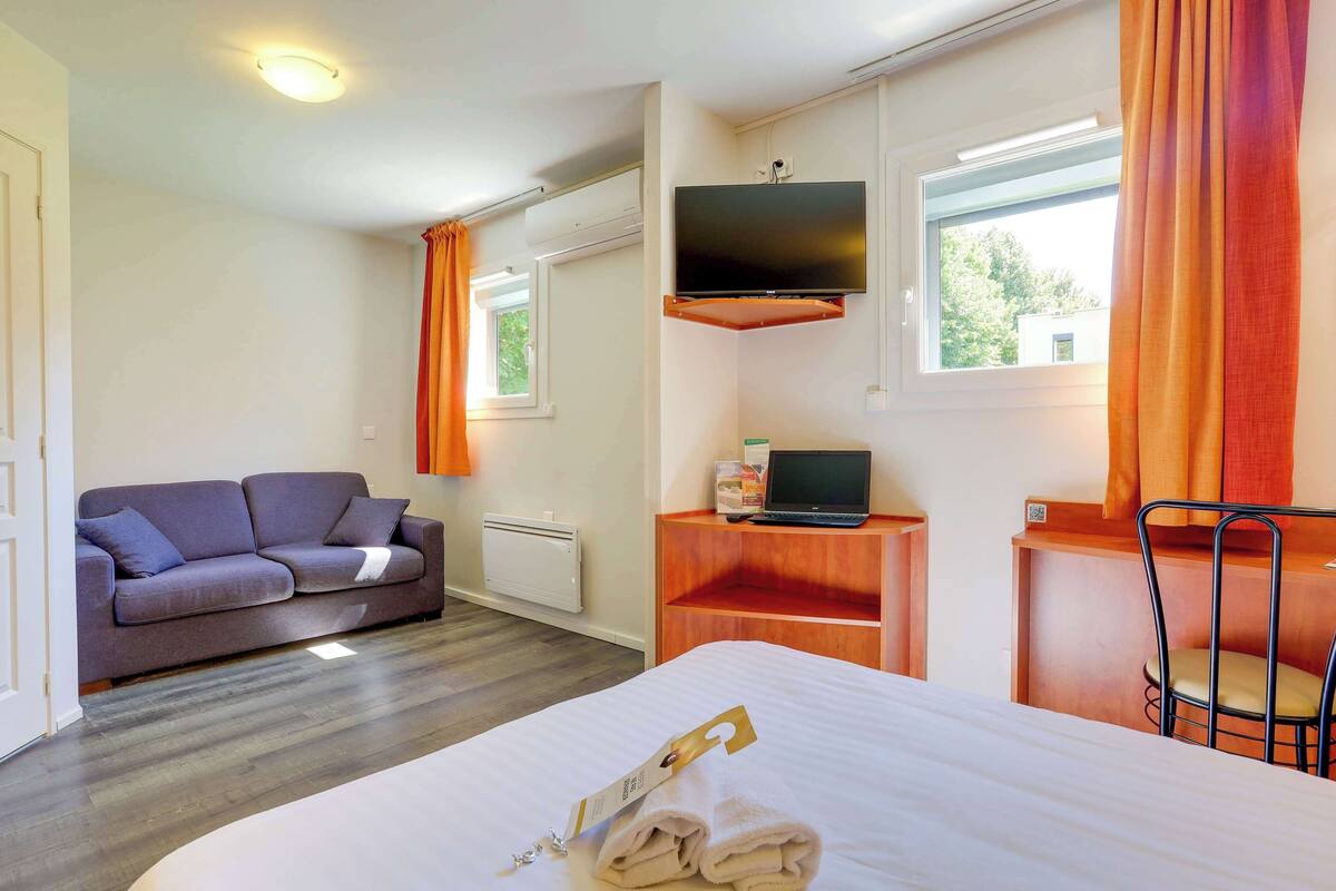 Habitación de hotel con cama, sofá, escritorio con computadora portátil, televisor montado en la pared y ventana con cortinas.