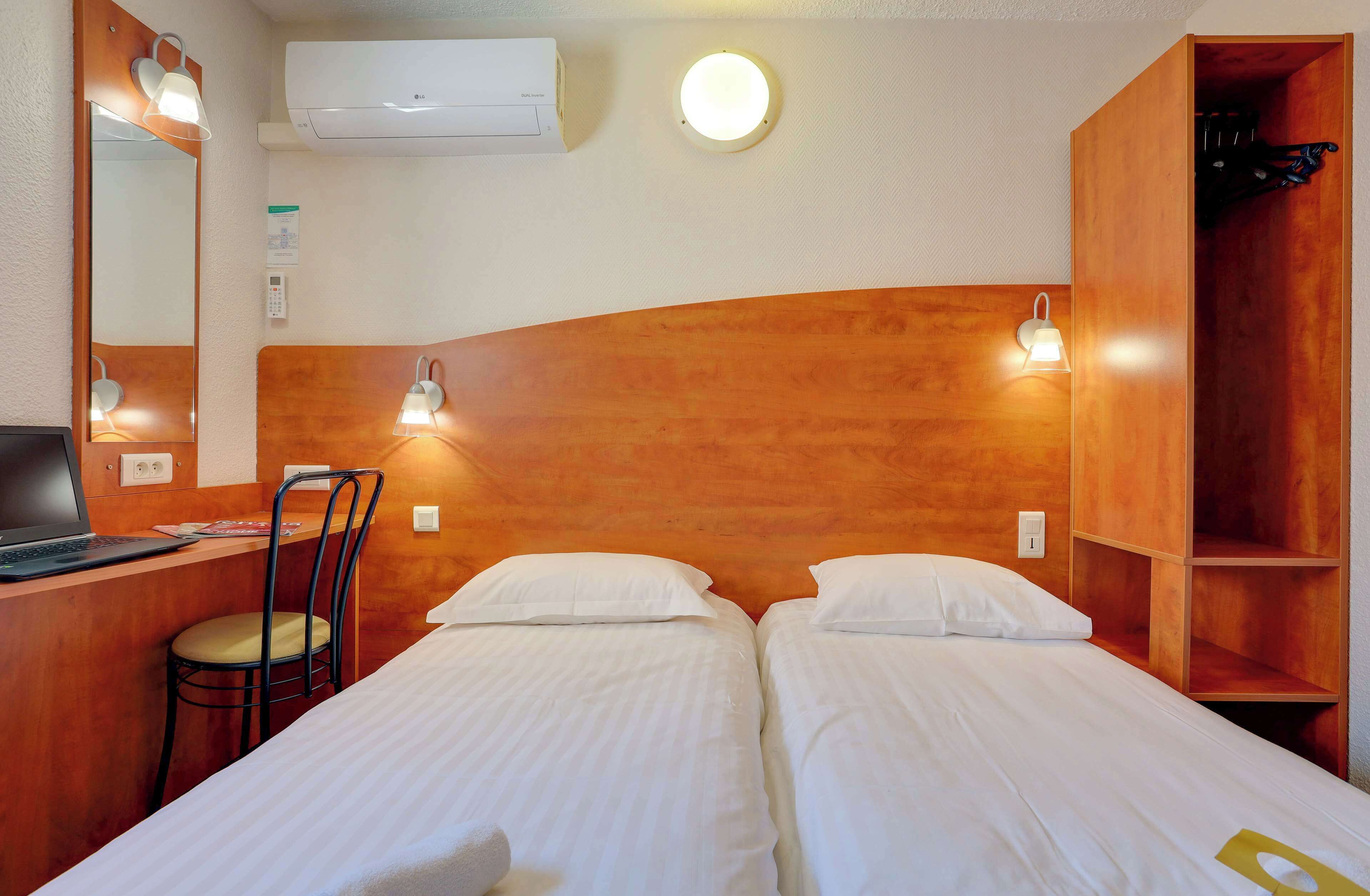 Hotellihuone, jossa on kaksi sänkyä, työpöytä kannettavalla tietokoneella, tuoli, peili ja vaatekaappi.