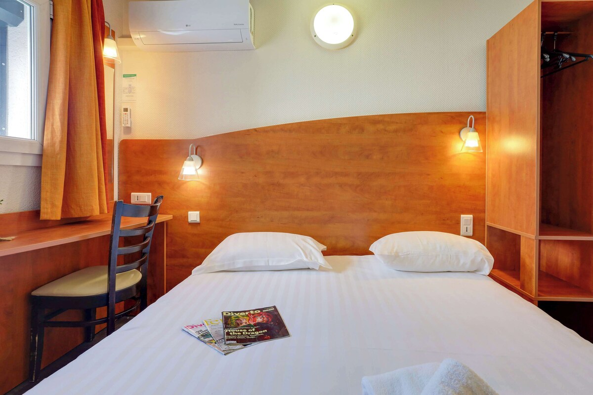 A hotel room with a wooden headboard, two white pillows, a wooden desk with a chair, a magazine on the bed, and a ceiling light.