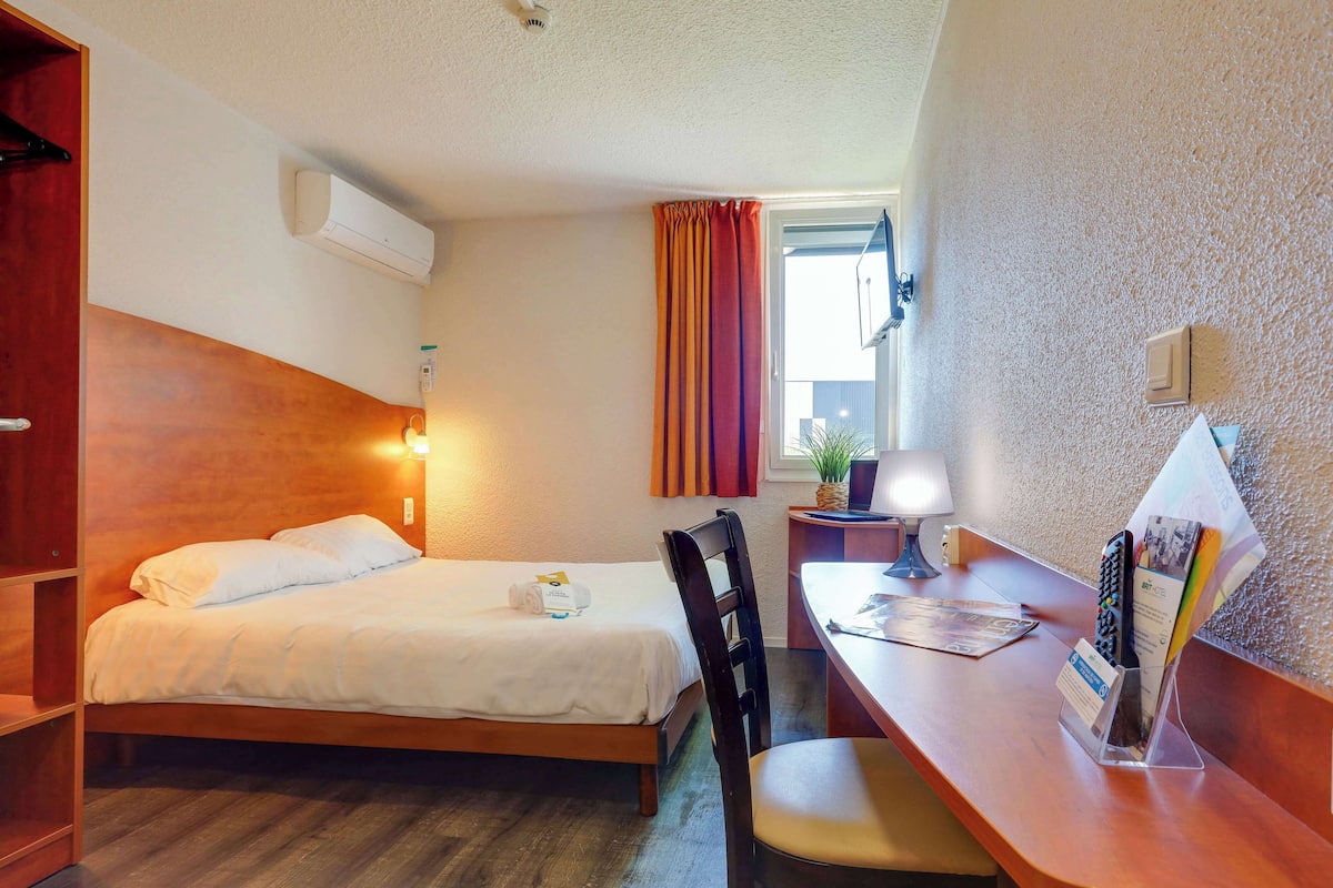 Una habitación de hotel compacta con una cama individual, un escritorio con silla, una mesita de noche con lámpara, una ventana con cortinas y un equipo de aire acondicionado.
