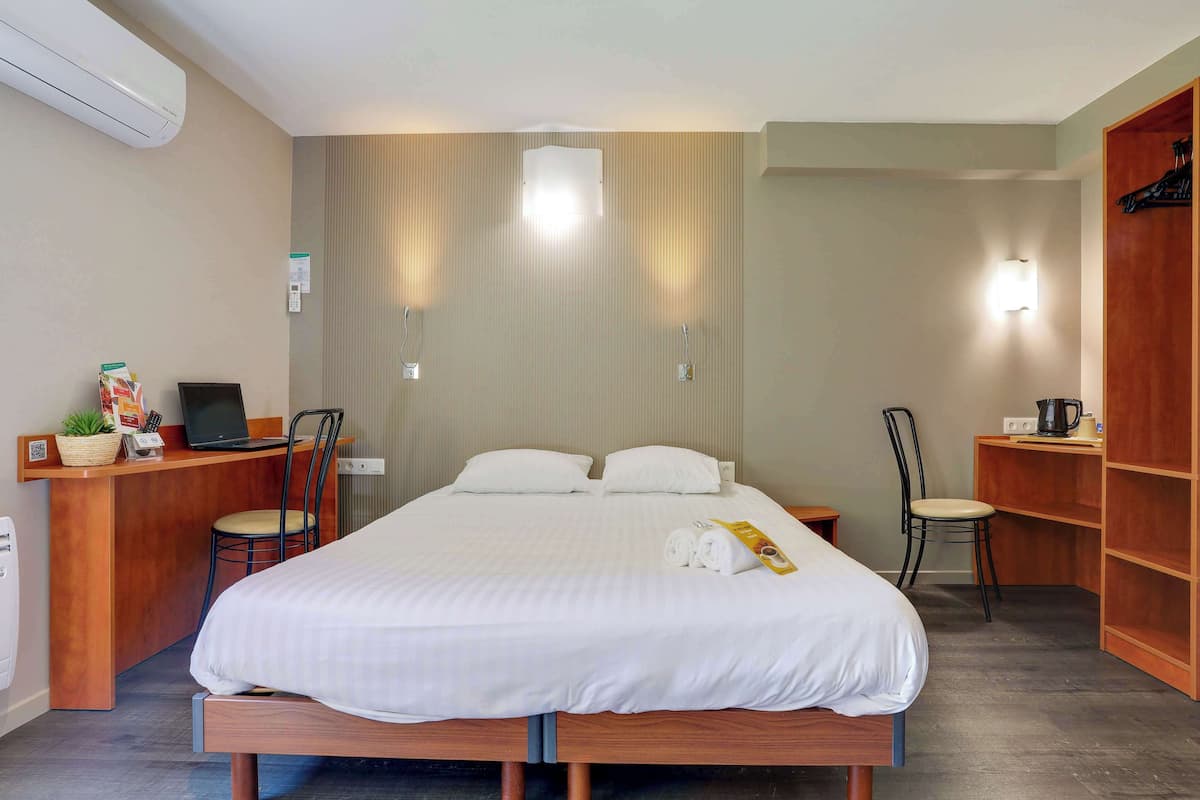 Habitación de hotel impecable con una cama grande, un escritorio con una computadora portátil, una silla y una estantería con un hervidor.