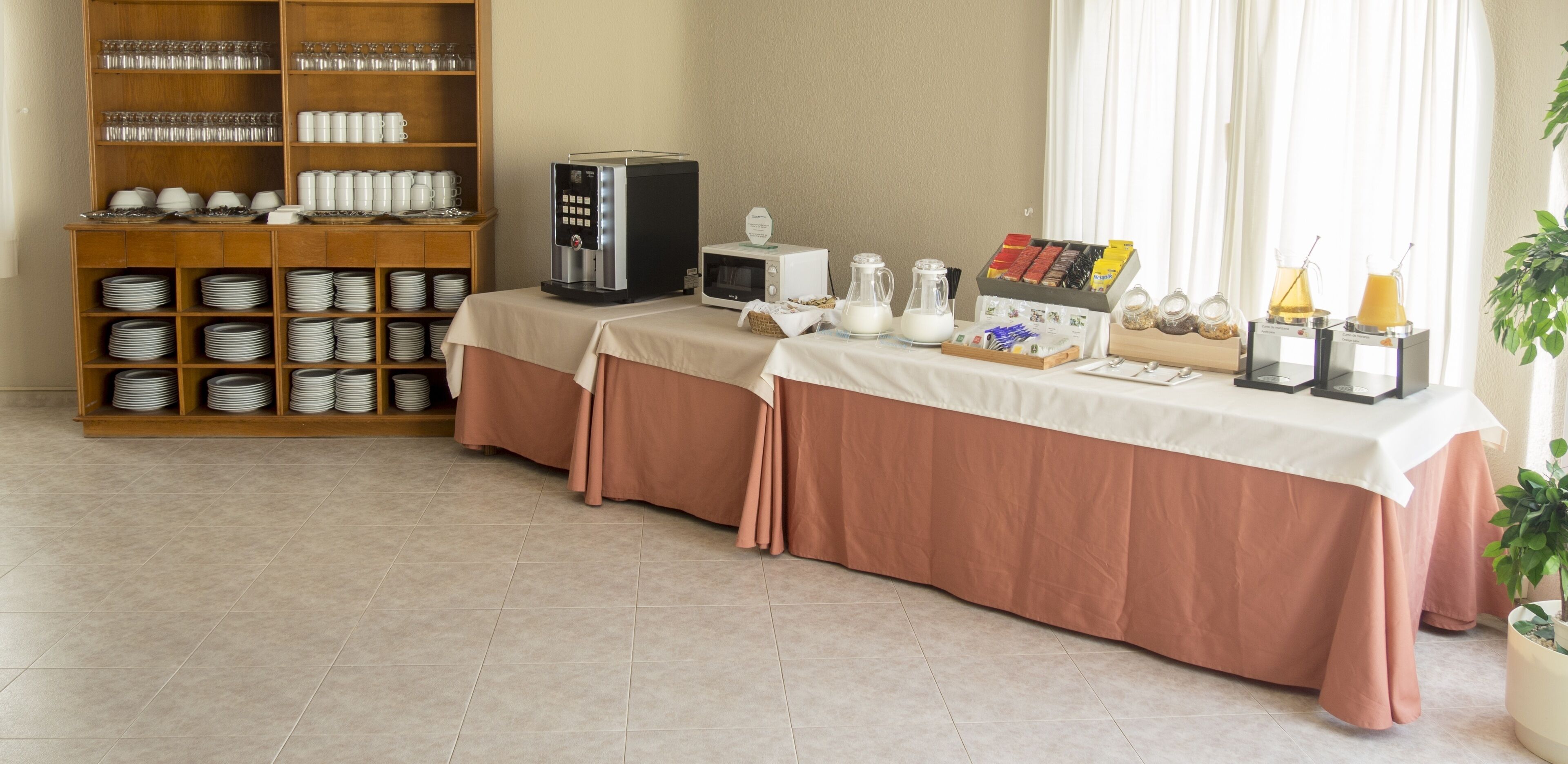 Montaje para desayuno con cafetera, microondas y diversos alimentos sobre una mesa, con una zona de bufé al fondo.