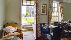 A dining area with a table set for two, a sofa with a floral cushion, and a view of a historic building through open doors. - The Ancient Gatehouse Hotel (Wells)