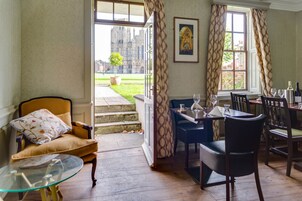 Un comedor con una mesa dispuesta para dos personas, un sofá con un cojín floral y vistas a un edificio histórico a través de puertas abiertas.