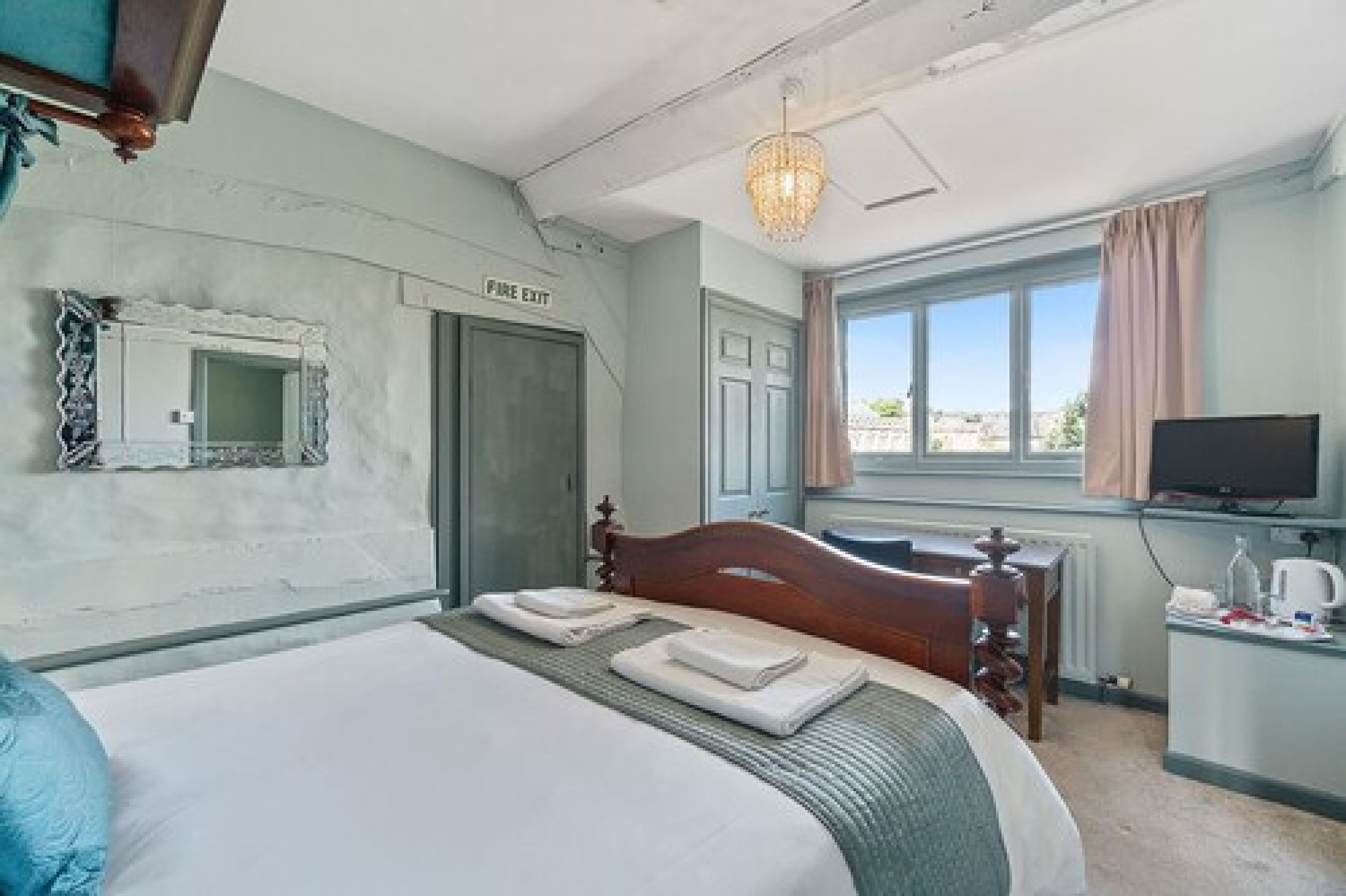A bedroom with a bed, a bedside table, a television, a window with curtains, and a 'Fire Exit' sign.