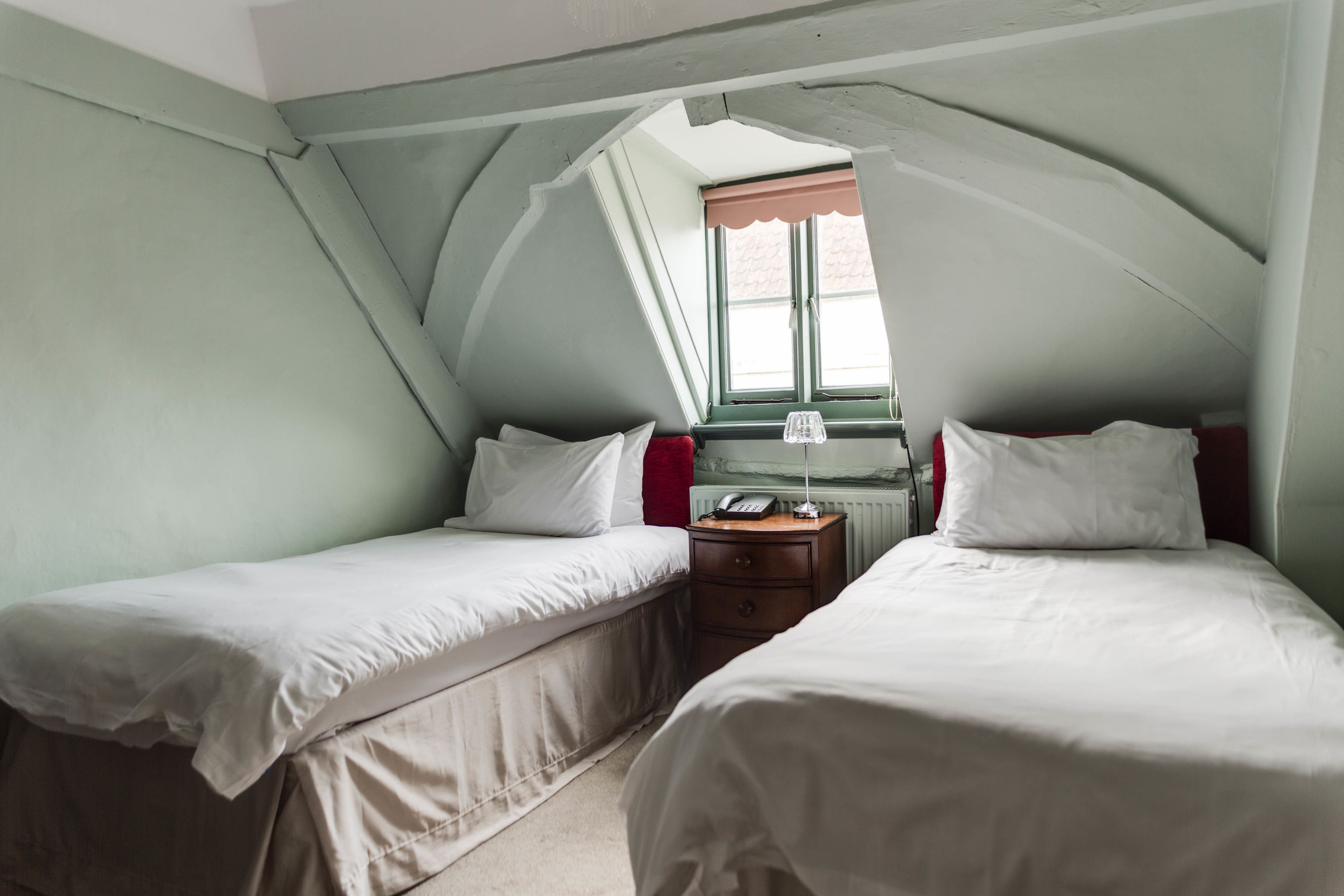 A small, cozy room with two single beds, a nightstand with a lamp, and a window with a view.