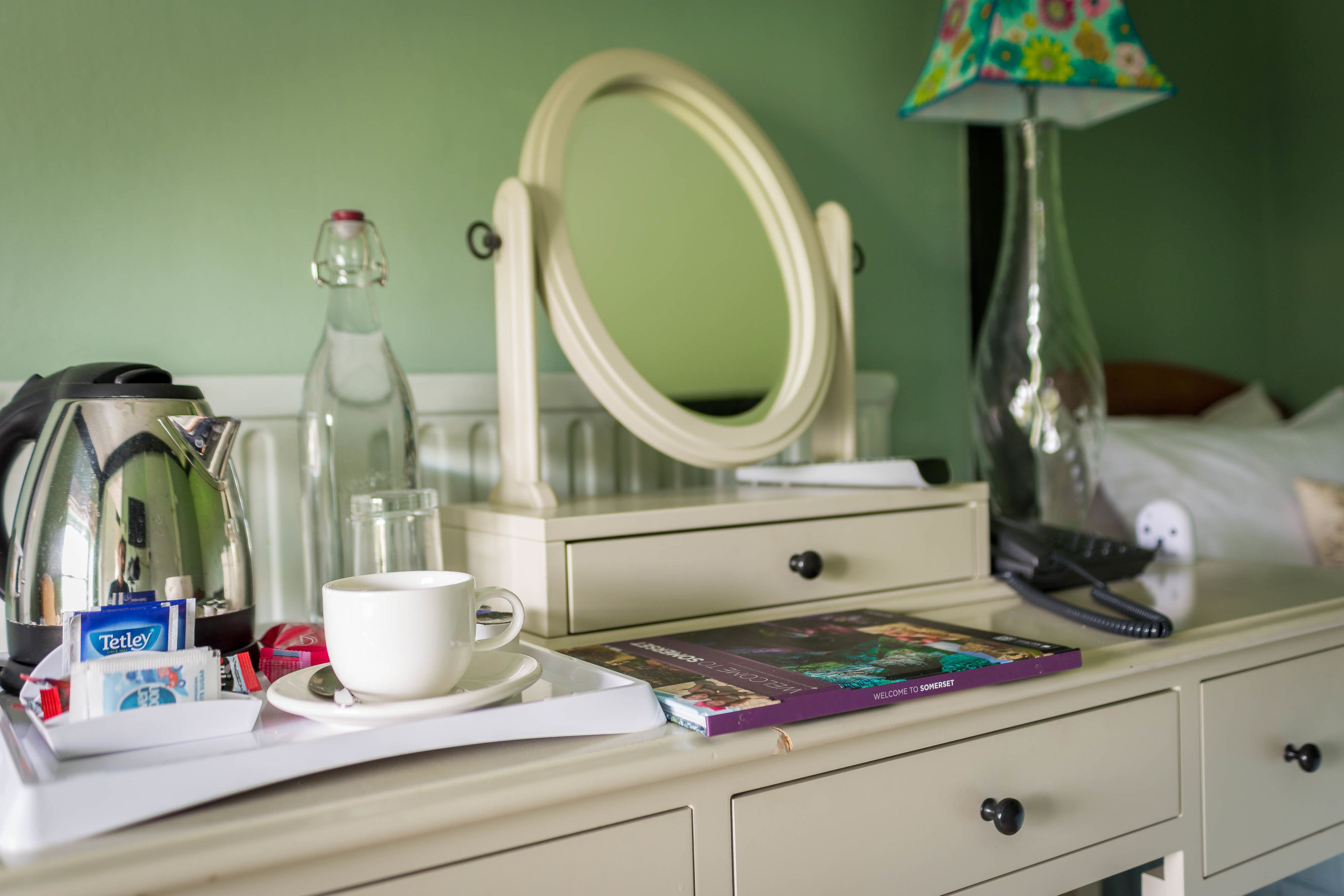 A hotel room with a white bedside table, a mirror, a lamp, a book, a cup, and a kettle.
