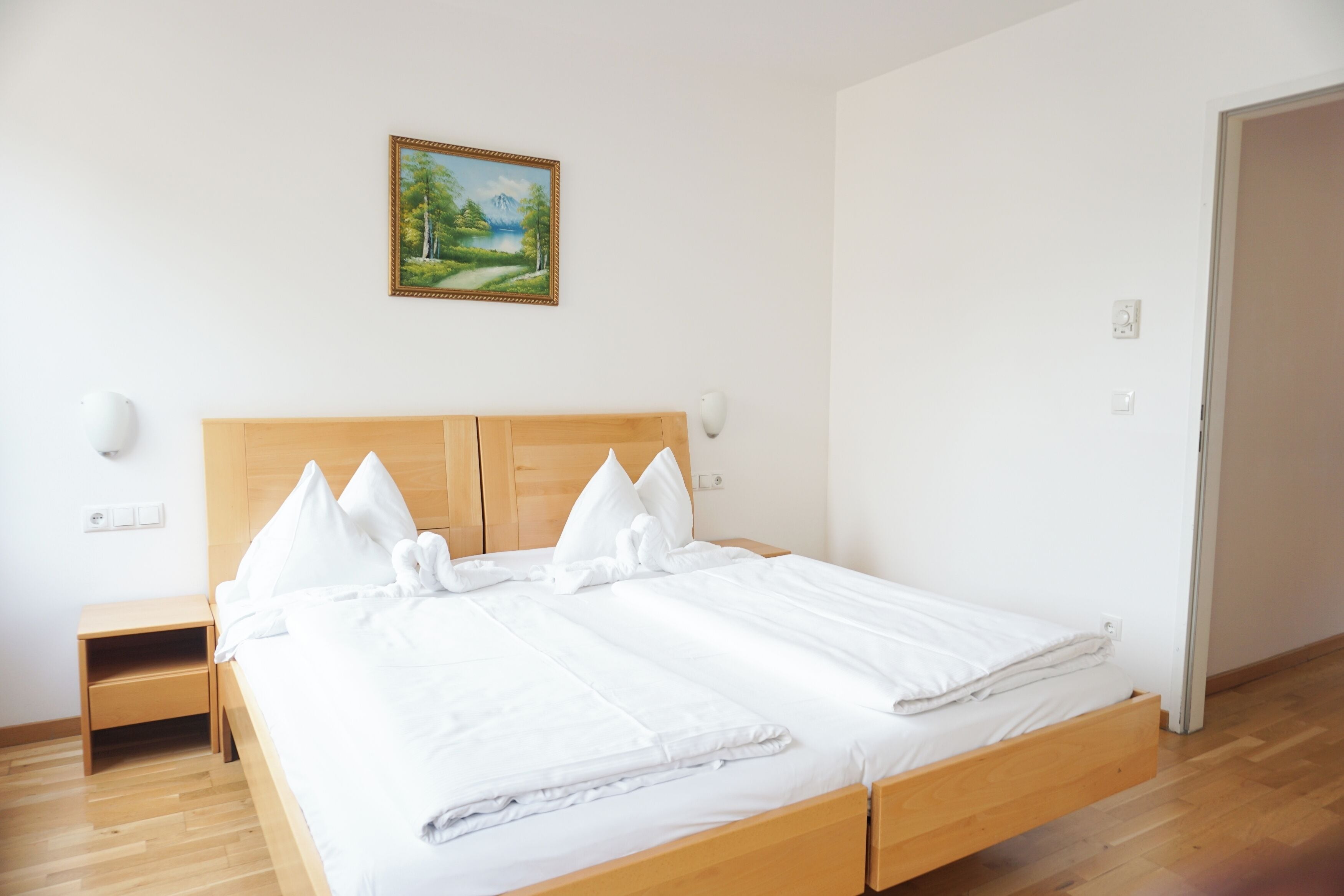 A neatly made bed with white linens, a wooden headboard, a nightstand with a lamp, and a painting of a landscape on the wall.