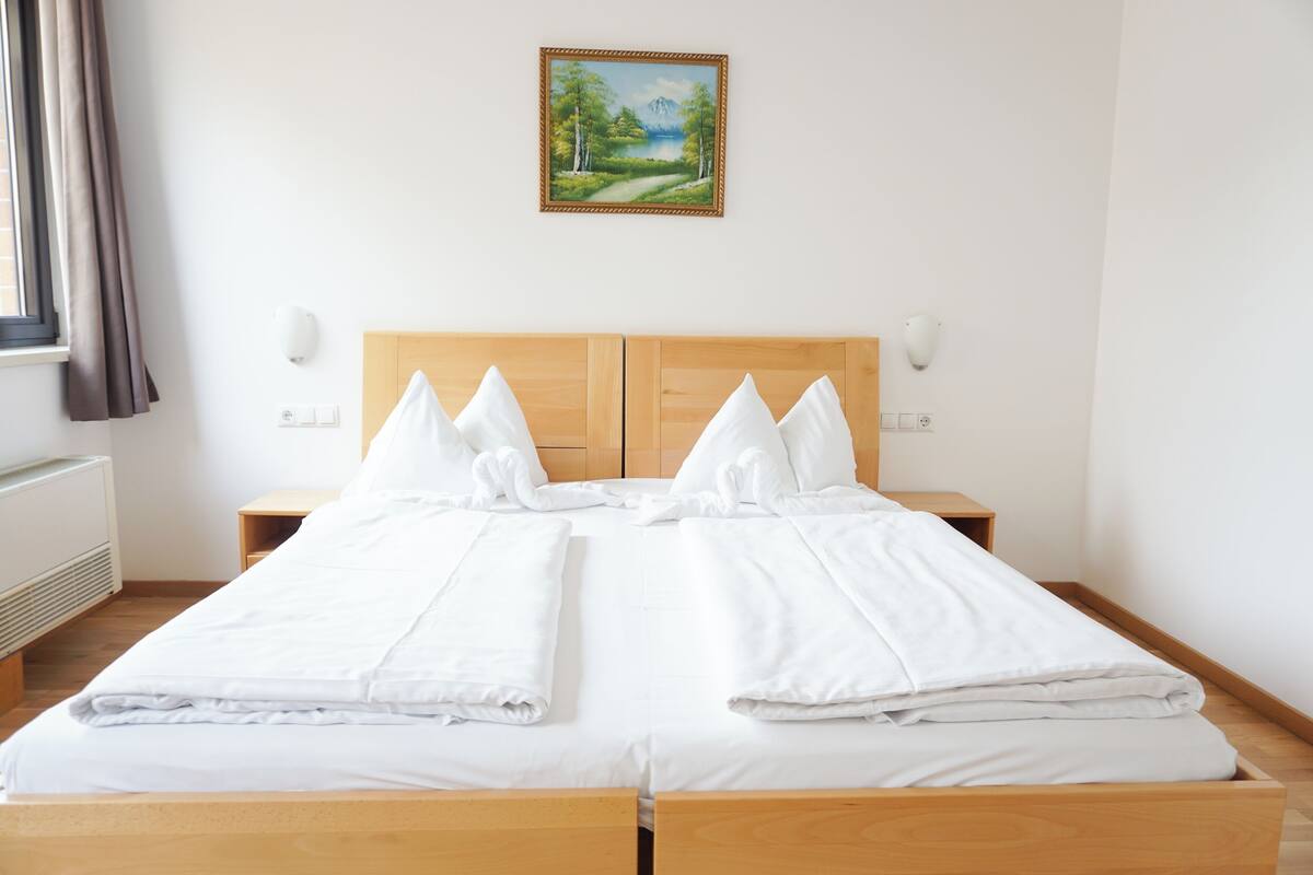 A double bed with white linens, a wooden headboard, bedside tables with lamps, and a painting of a landscape on the wall.