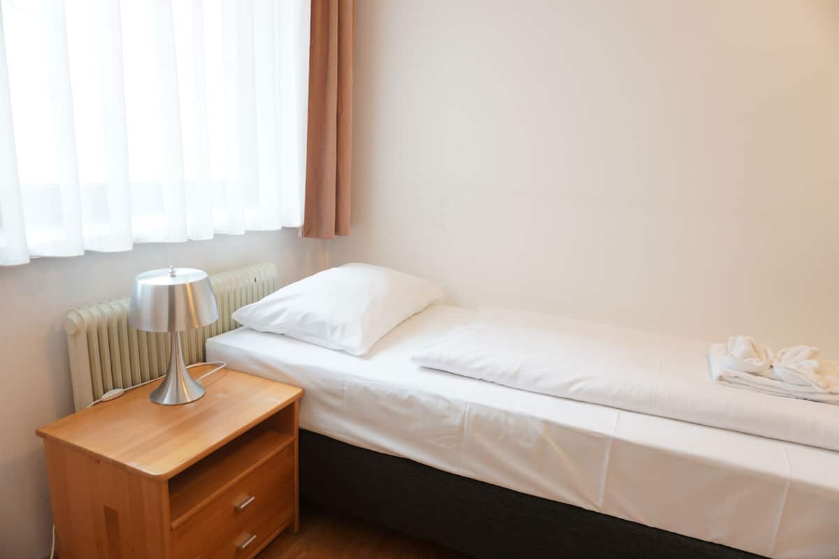 A neatly made bed with white bedding, a bedside table with a lamp, and a radiator.