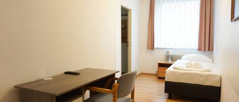 A bedroom with a wooden desk, a chair, a bed with a white duvet, and a window with curtains.