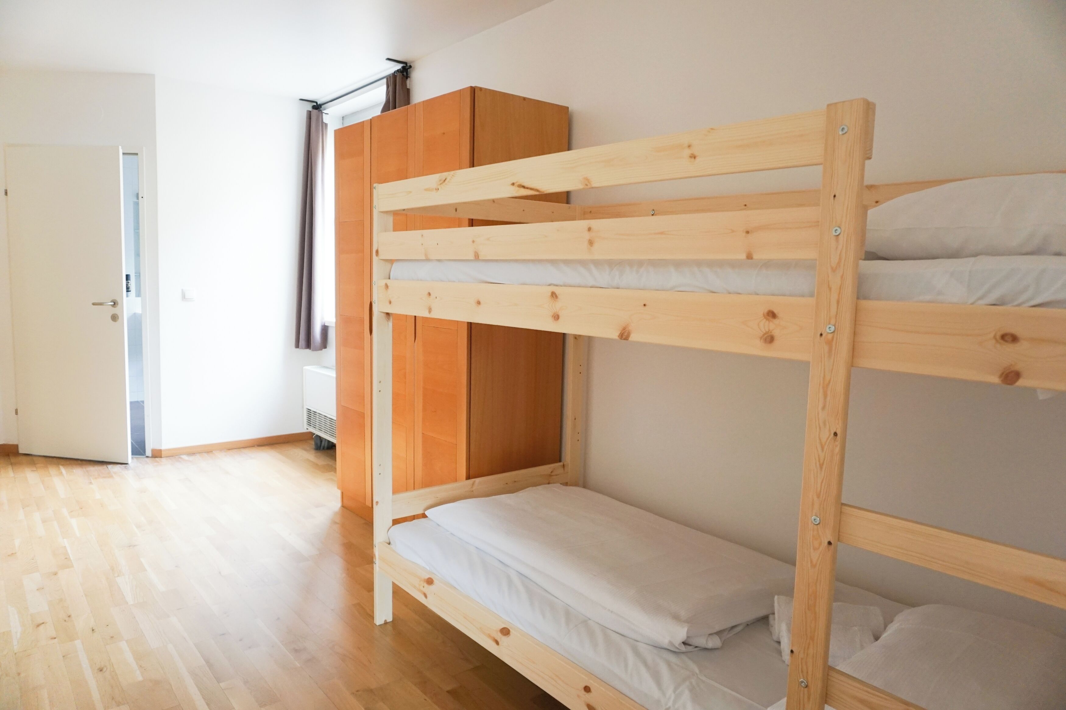 Una cama litera con ropa de cama blanca, un armario de madera y una puerta que da a otra habitación.