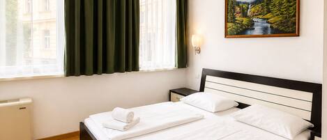 A double bed with white linens and towels, a nightstand, a wall-mounted lamp, a painting of a landscape, and a window with curtains.