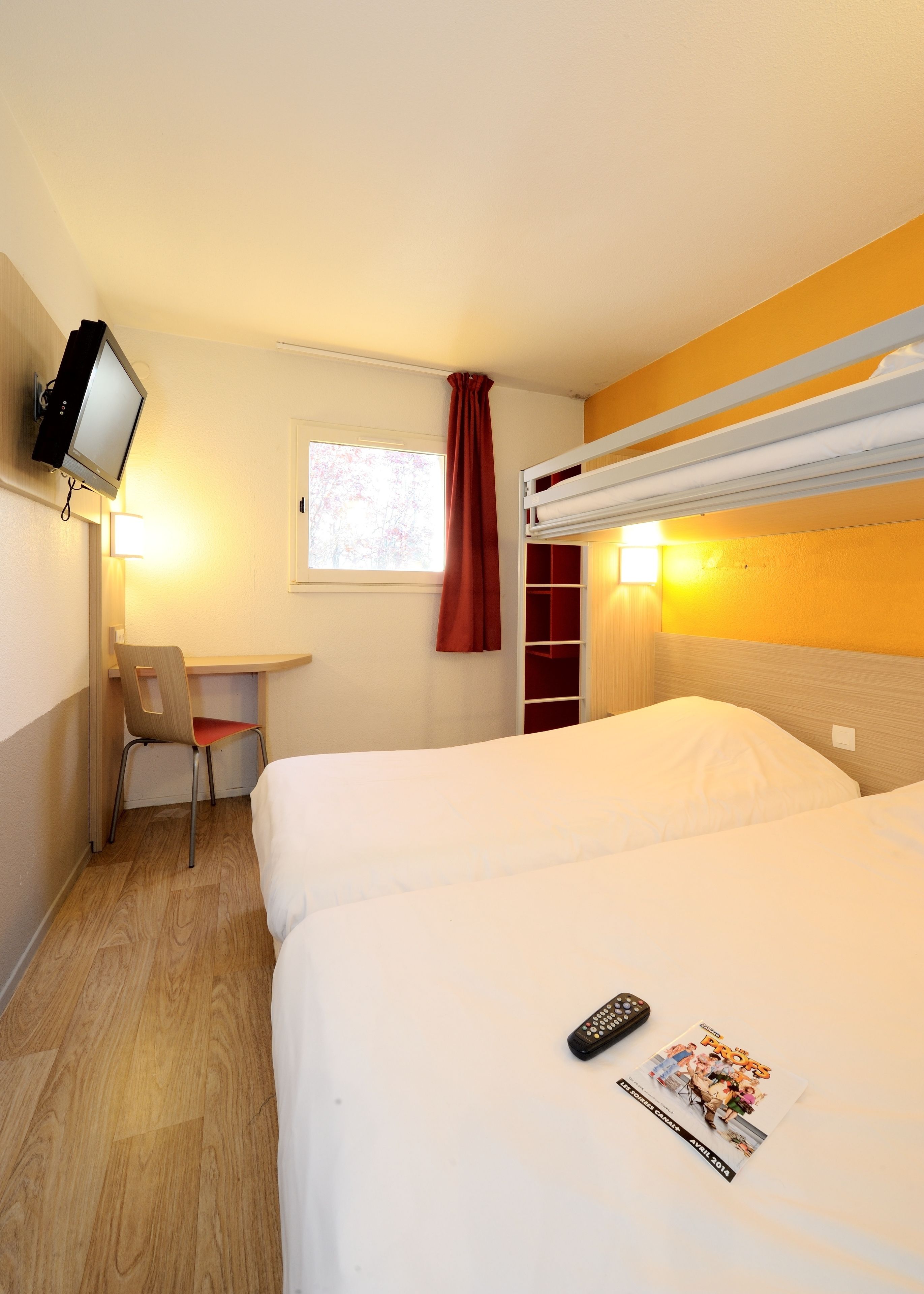 Une chambre d’hôtel avec un lit superposé, un bureau avec une chaise, un téléviseur fixé au mur et une fenêtre avec un rideau rouge.