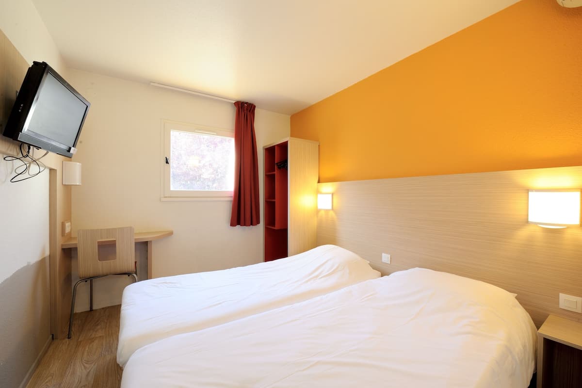 Hotellihuone, jossa on kaksi sänkyä, seinälle asennettu televisio, pieni työpöytä ja ikkuna, jossa on verhot.